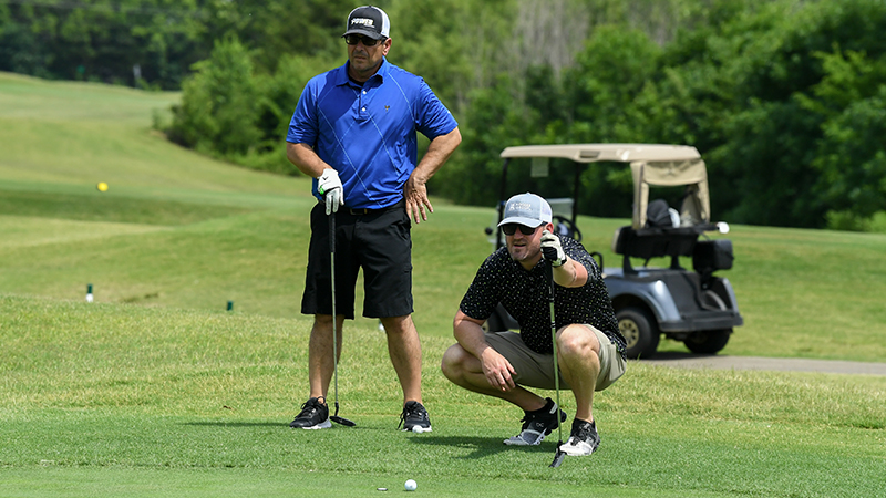 A man in a blue shirt and sunglasses stands with a golf club, while another man in a patterned shirt crouches beside a golf ball on a golf course